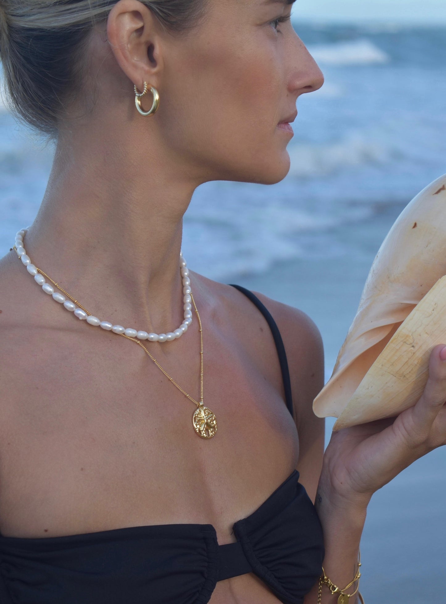 Model wearing short pearl necklace layered with Serene Scenes The Label 18K gold-filled Sand Dollar Necklace. Gold chain with circular pendant featuring starfish and textured design.